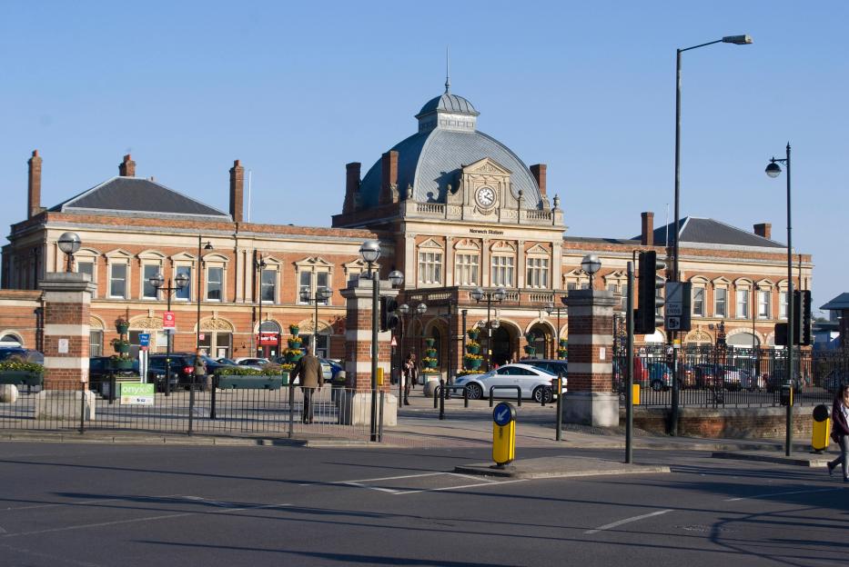 Norwich train station, a large brick building with a domed roof and clock tower, seen from across a street with traffic and pedestrians.