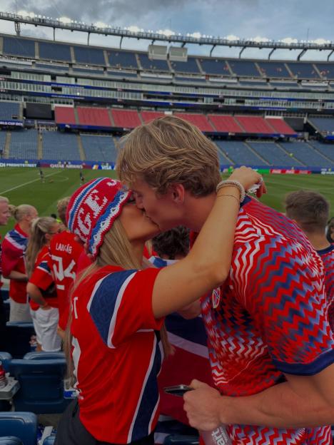 A couple in Norwegian flag-themed attire kiss in a stadium.