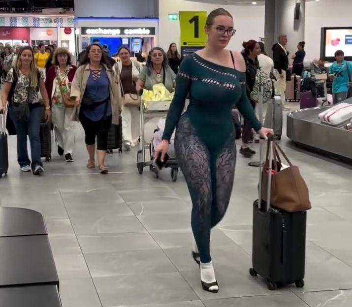 A woman in a revealing, form-fitting green and black outfit walking through an airport with a rolling suitcase.