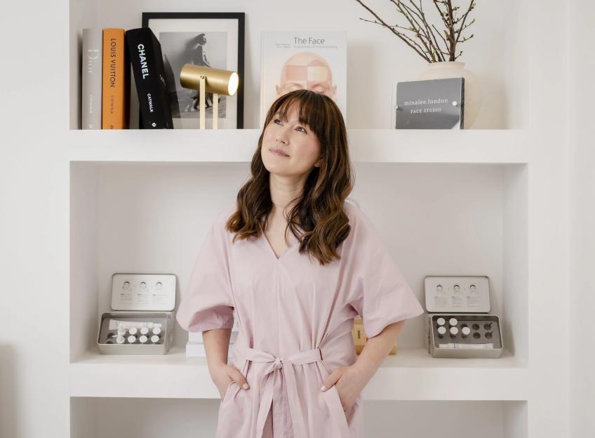 Mina Lee in a pink dress, standing in front of shelves with books and cosmetic products.