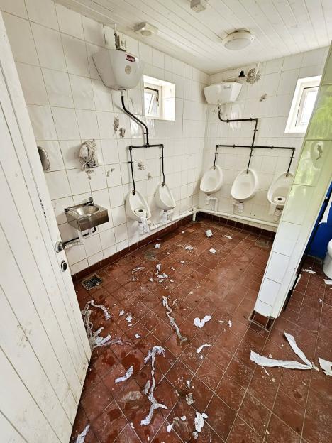 A dirty, vandalized men's public restroom with paper and debris on the floor.