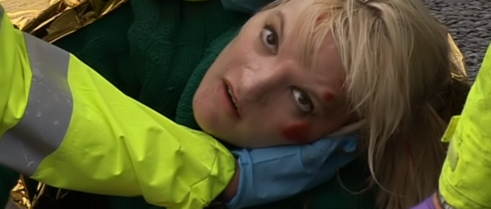 Ursula Eriksson with blonde hair, red marks on her face, being assisted by someone in a yellow jacket and blue gloves.