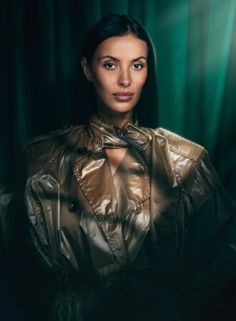 Maya Jama wearing a shiny trench coat against a dark green curtain background.