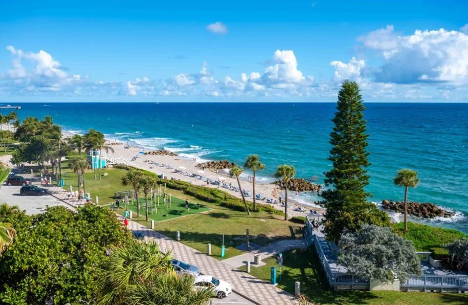 High-angle aerial view of Deerfield Beach in Florida with turquoise water, palm trees, people, and a blue sky with clouds.
