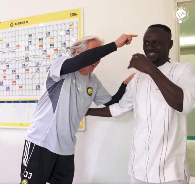 A man in a grey shirt laughing and pointing at another man in a white shirt, who is also smiling, with a calendar board in the background.
