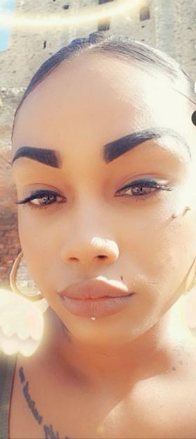 Close-up of a woman's face with light brown skin, dark hair pulled back, and dark eyebrows, wearing a small stud below her lower lip.