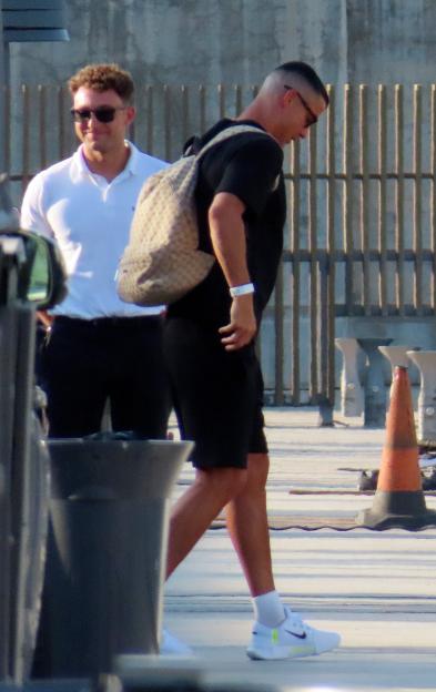 Cristiano Ronaldo walking with a Gucci backpack and white wristband, while a man in a white polo shirt and sunglasses smiles behind him.