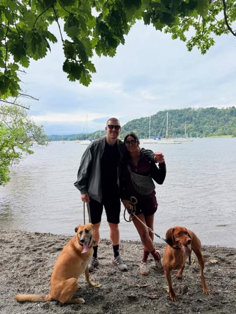 Karen Hauer and her boyfriend standing with two dogs by a lake with sailboats.