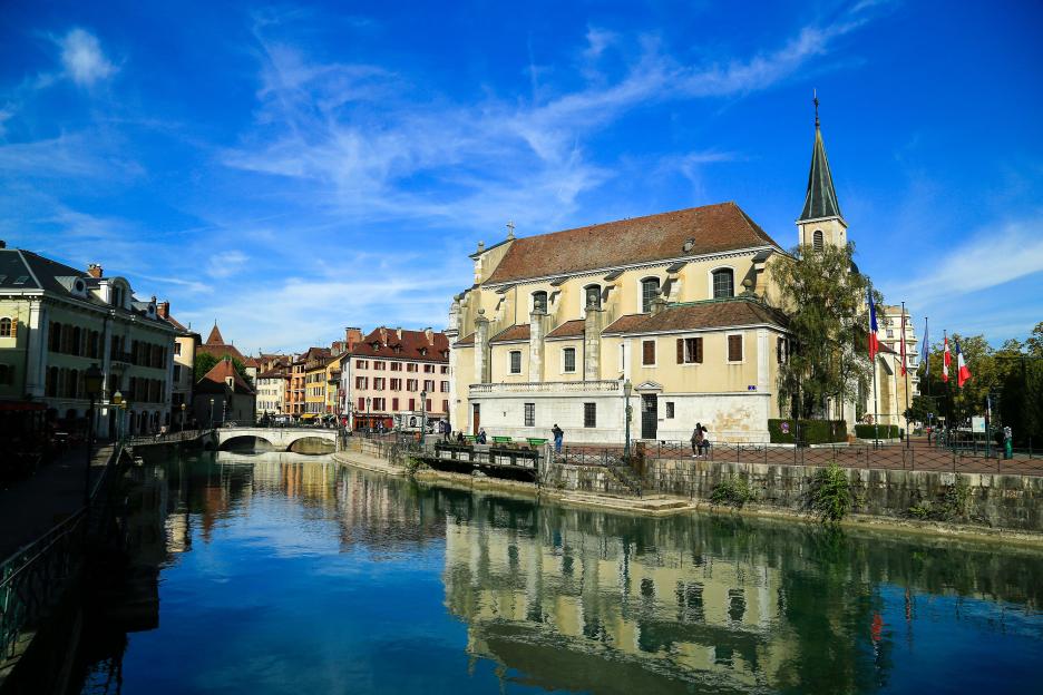 View of Annecy, France, with a calm canal reflecting the colorful buildings and a church with a tall steeple under a clear blue sky.