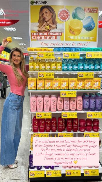 A woman in a pink top and jeans poses next to a Garnier display in a store, holding up a Garnier product.