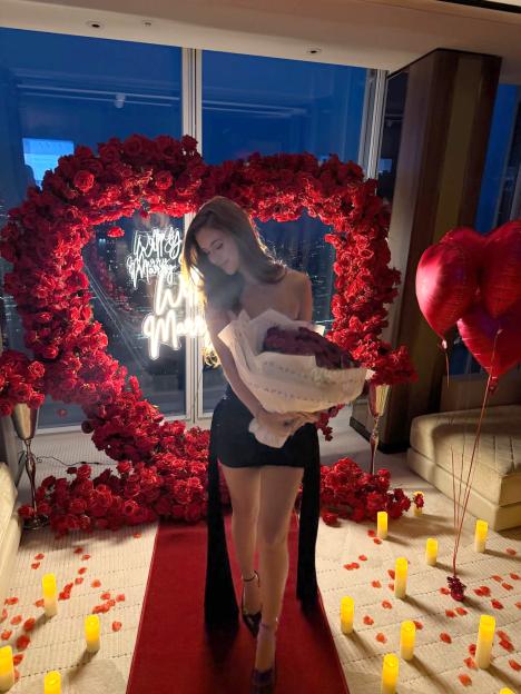 Amanda Araujo holding a bouquet of red roses, standing on a red carpet with candles and rose petals, in front of a heart-shaped rose arch and a "Will You Marry Me" neon sign.