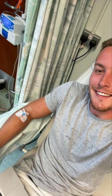 A smiling man with an IV drip in his arm is sitting in a hospital room.