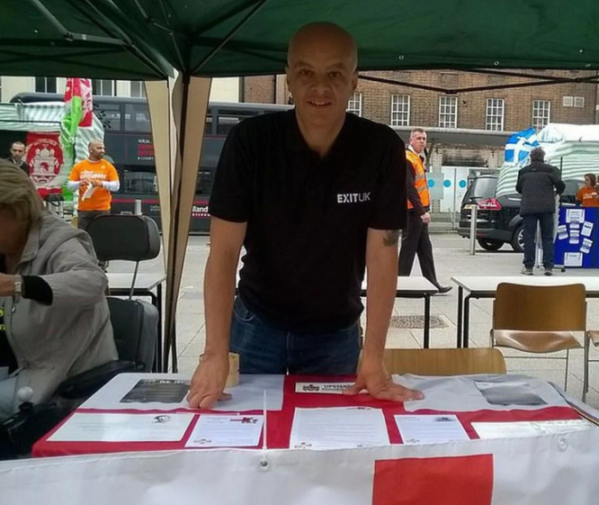 Man in an "EXITUK" shirt standing behind a table with an England flag tablecloth.
