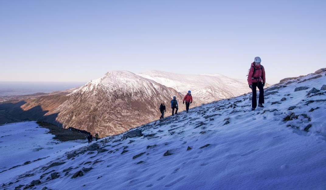 A group of five hikers ascending a snowy mountain slope with a vast mountain range in the background.