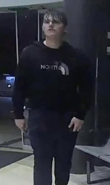 CCTV image of a young male with dark hair wearing a black hoodie and black trousers.