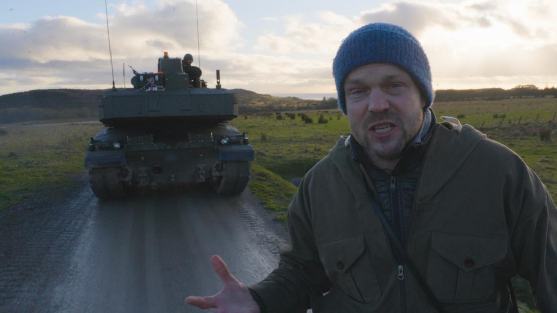 A man in a blue knit hat speaks to the camera with a tank behind him on a road.
