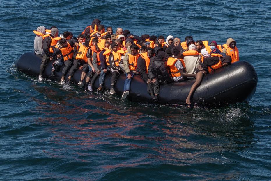 An inflatable boat crowded with refugees wearing orange life jackets on the sea.
