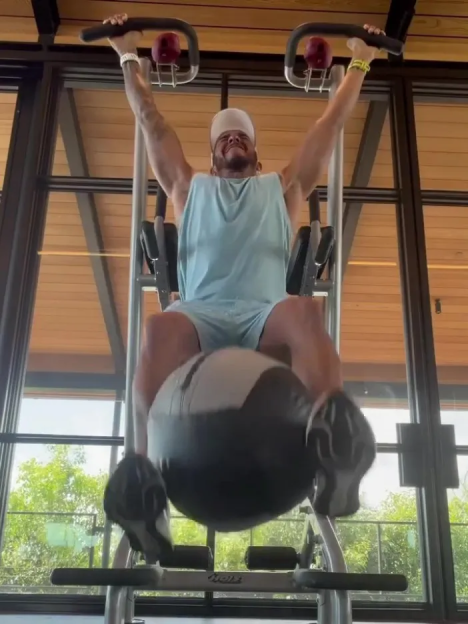 Man exercising on a hanging knee-raise machine with a weighted ball between his legs.