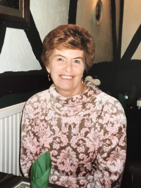 June Buffham, a smiling woman with short reddish-brown hair, wearing a patterned pink and brown top and a gold chain.
