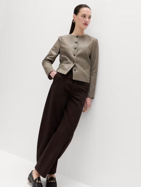 A woman models a faux leather collarless jacket with brown trousers and loafers.