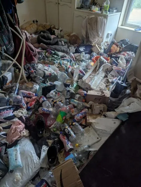 A room littered with rubbish, including plastic bottles, cardboard boxes, and clothes, with a cat sitting amidst the mess.