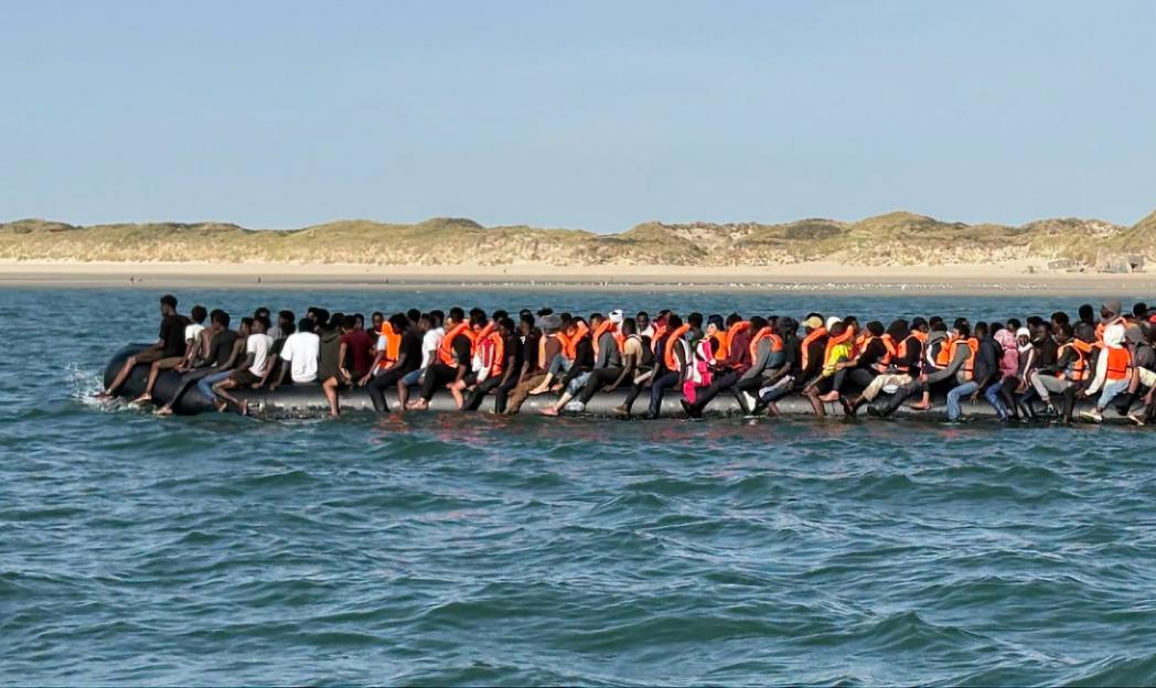 A large group of people packed into a single inflatable boat on rough water, with a sandy beach and dunes in the background.