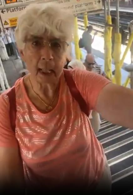 An elderly woman with white hair and glasses, wearing a peach-colored shirt, filmed herself in front of a train station platform sign.