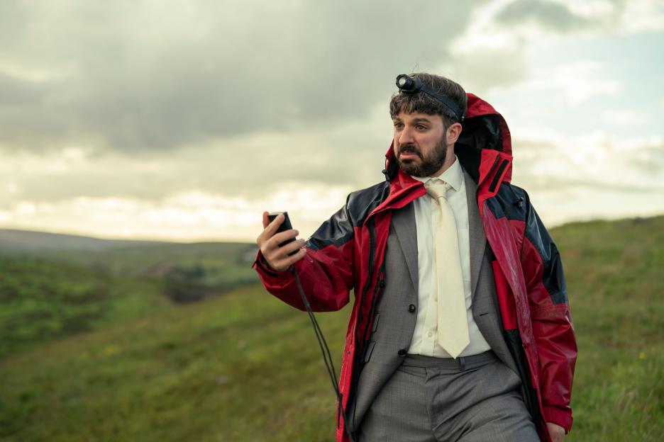 Simon Bird as David, wearing a headlamp and red jacket over a suit, holding a device in a field.
