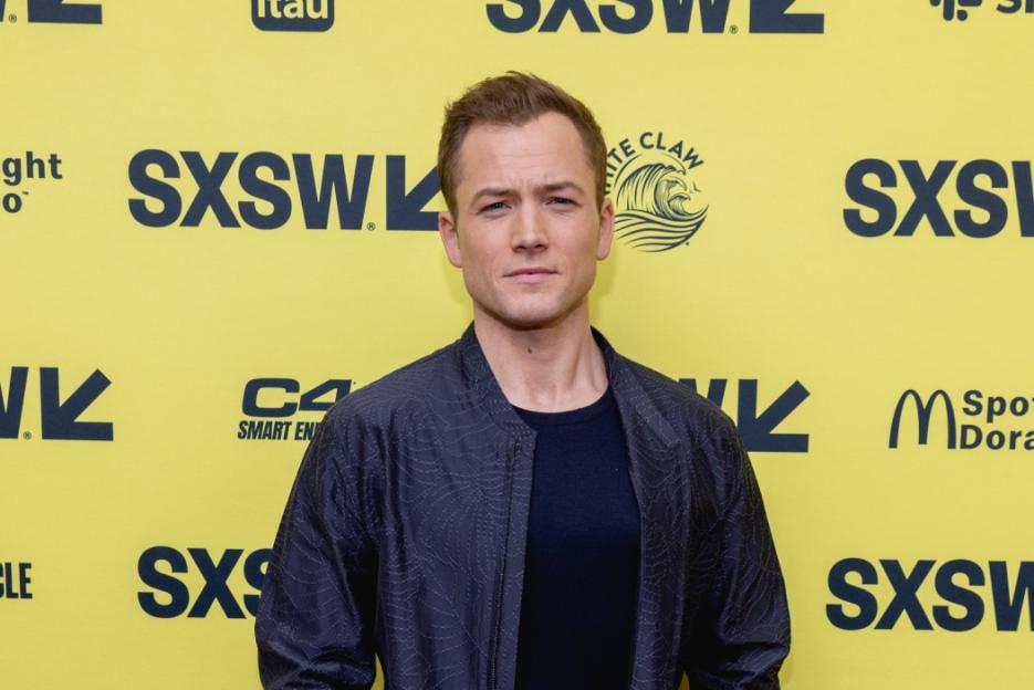 Taron Egerton posing on a yellow backdrop with various logos for the 2023 SXSW Conference and Festival.