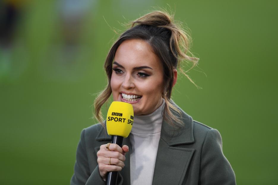 Kelly Somers, BBC Sport presenter, holding a yellow BBC Sport microphone and smiling pitchside.