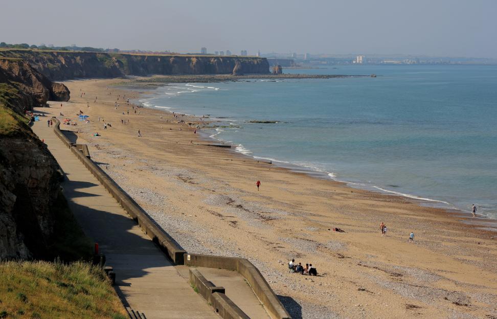 Seaham Beach in County Durham, with a paved walkway curving along the cliffside next to the sandy shore, where people are walking and relaxing, and the ocean stretches out to the horizon with a distant city skyline.