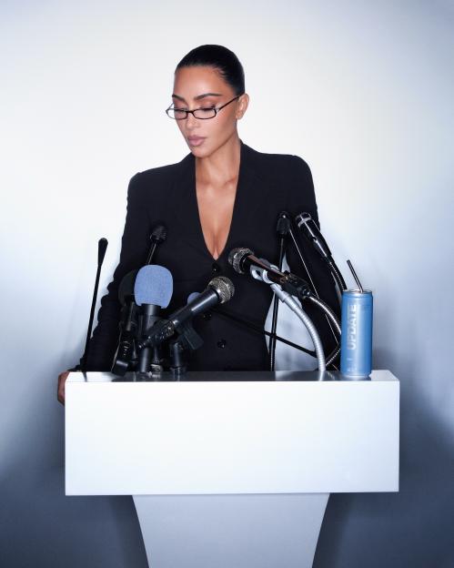 Kim Kardashian in a black suit with a plunging neckline, wearing glasses, standing behind a podium with microphones and a blue "UPDATE" drink can.