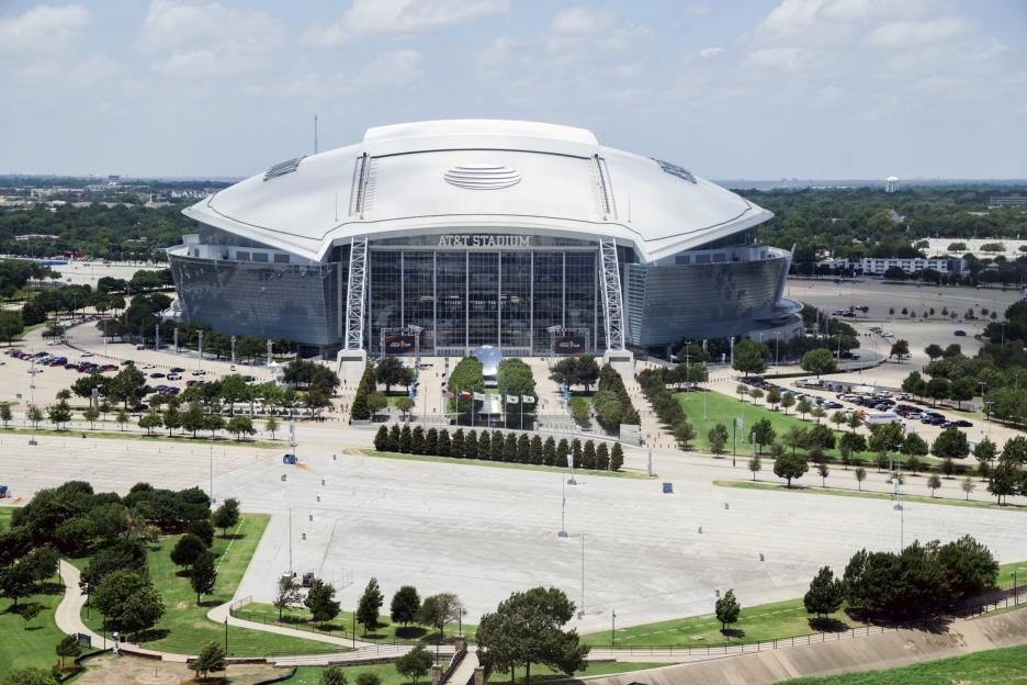 AT&T Stadium, an oval-shaped building, with 'AT&T STADIUM' written in large black letters across the entrance, with several parking lots surrounding it.