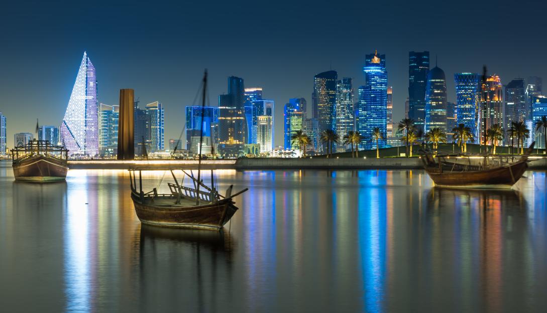 Illuminated city at waterfront in Doha, Qatar, with two traditional boats in the foreground.