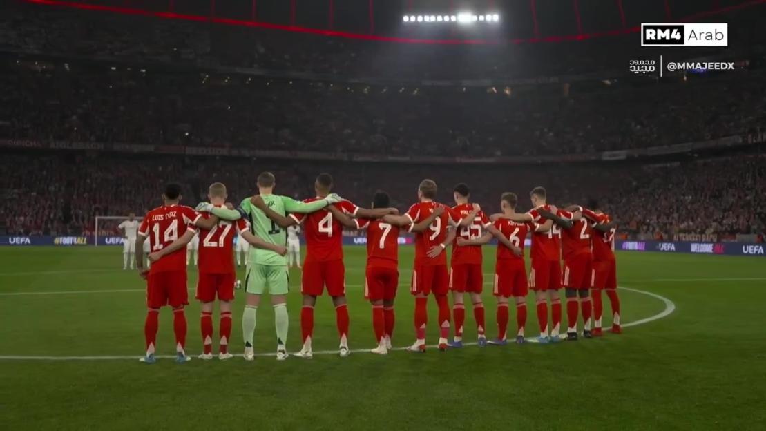 A row of Bayern Munich football players with their arms around each other, facing away from the camera, on a green field in front of a stadium full of spectators.