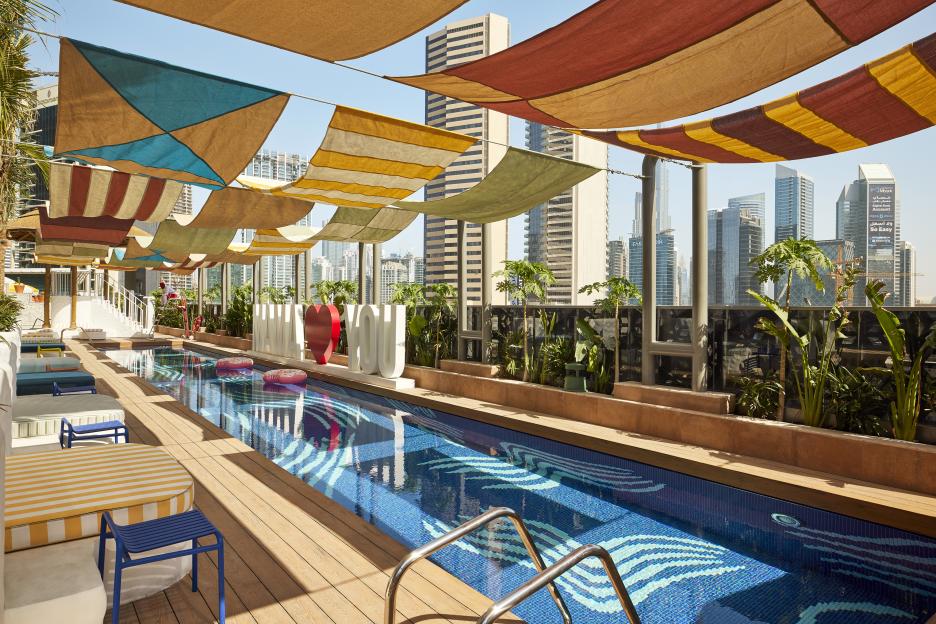 Rooftop swimming pool with lounge chairs and shaded by colorful awnings, overlooking the Dubai skyline.