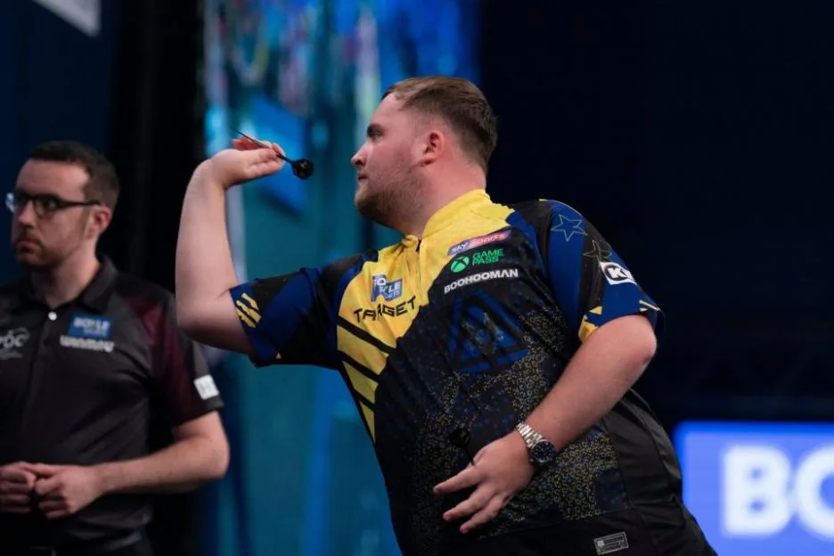 A darts player in a yellow and blue shirt aims to throw a dart at the World Grand Prix, with an official watching in the background.