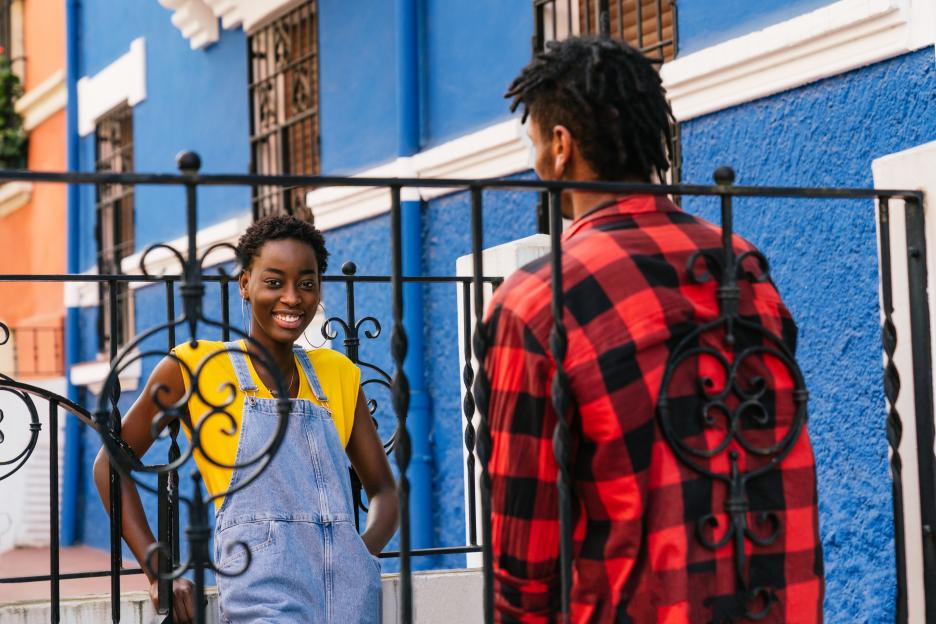 Young woman in yellow shirt and denim overalls laughing through a wrought-iron fence.