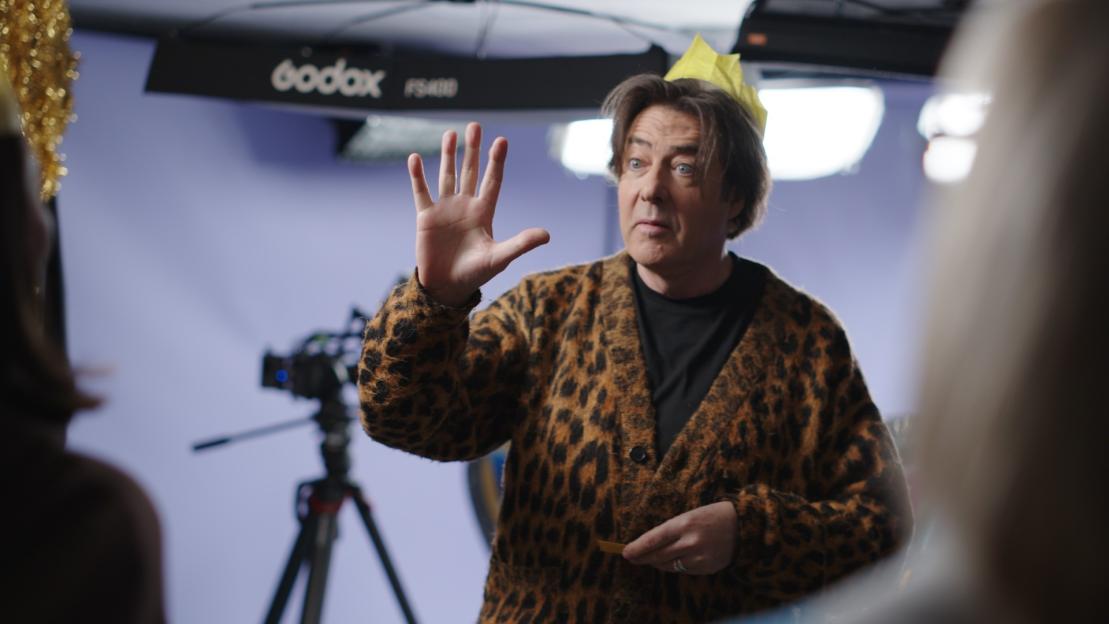 A man in a leopard print cardigan and a paper crown raises his hand, appearing to talk to someone off-camera.