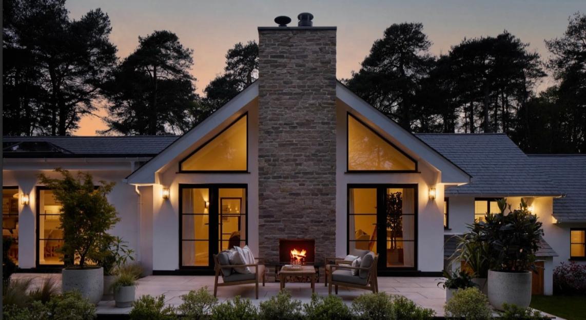 Modern house with a large stone chimney and outdoor seating area with a lit fireplace at dusk.