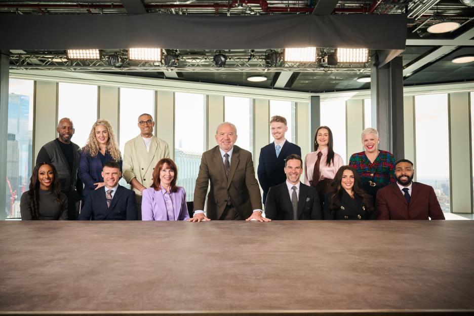 A group photo of 12 celebrities and Lord Sugar, the host of "The Celebrity Apprentice," in an office with large windows overlooking a city.