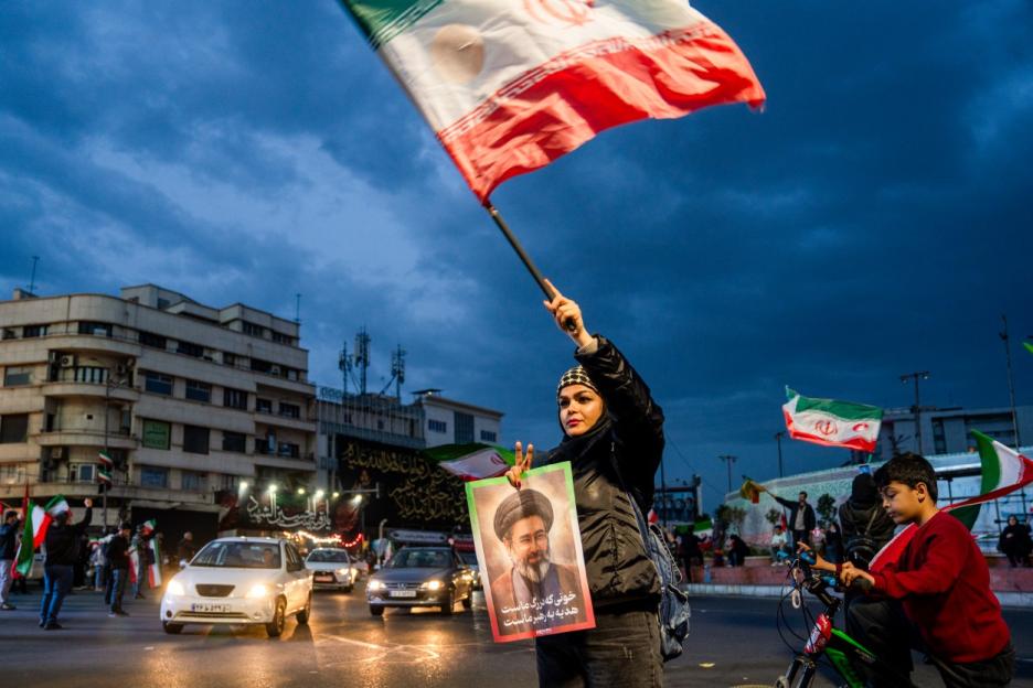 A person holding an Iranian flag and a poster of an Iranian leader, with a street scene in the background.