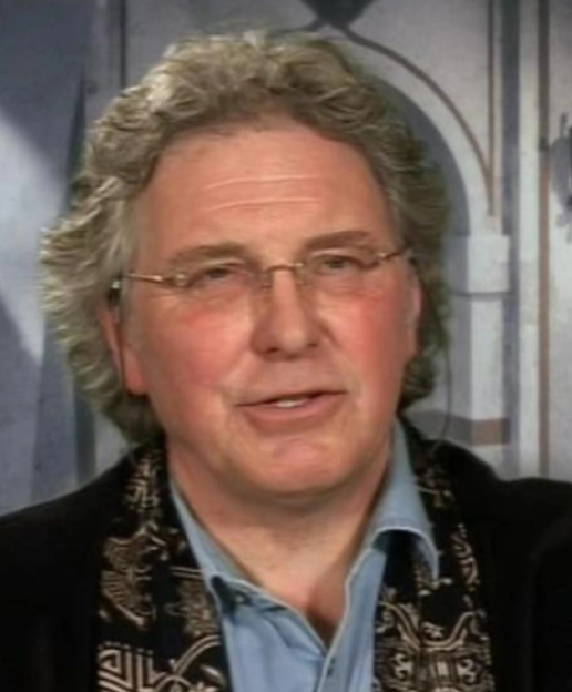 Headshot of Pual Seed, director, wearing glasses, a blue shirt, and a black patterned scarf.