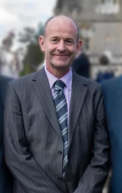 Jamie Gawler, a bald man in a pinstripe suit, purple striped shirt, and patterned tie, smiles at the camera.