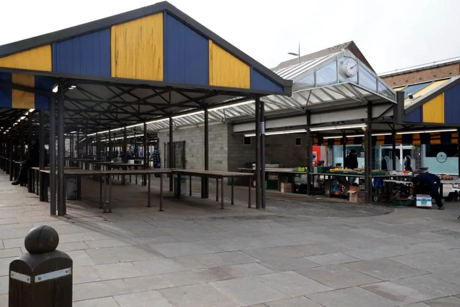 An outdoor market with empty stalls under blue and yellow awnings, and a few vendors setting up in the background.