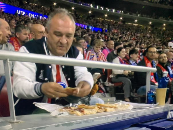 A man in a bomber jacket with a UEFA Champions League logo sitting in a stadium and eating a sandwich from a foil-wrapped tray of sandwiches.
