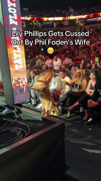 Video capture of a woman in a beige dress arguing with another woman in a black top, with text overlay "Lily Phillips Gets Cussed Out By Phil Foden's Wife".