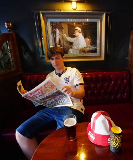 Ed Southgate, a Sun reporter, at the Sir Winston public house in Oslo, reading a newspaper, wearing an England football shirt.