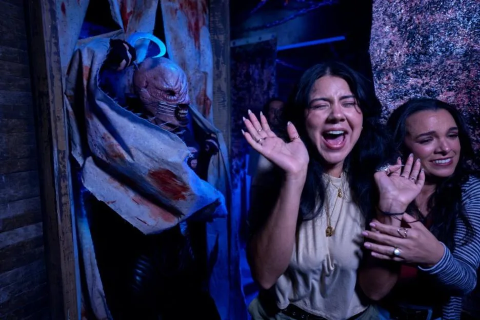 Two women react in fear to a terrifying monster holding a hook in a haunted maze.