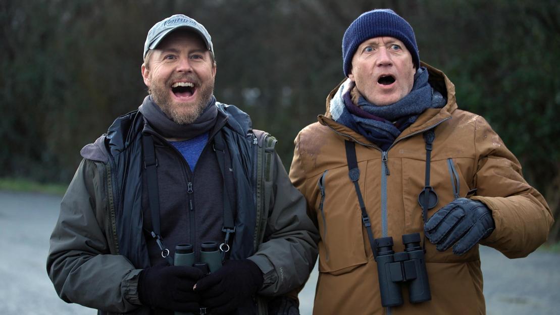 Two men in winter gear with binoculars around their necks, one smiling and the other with an open-mouthed, surprised expression.